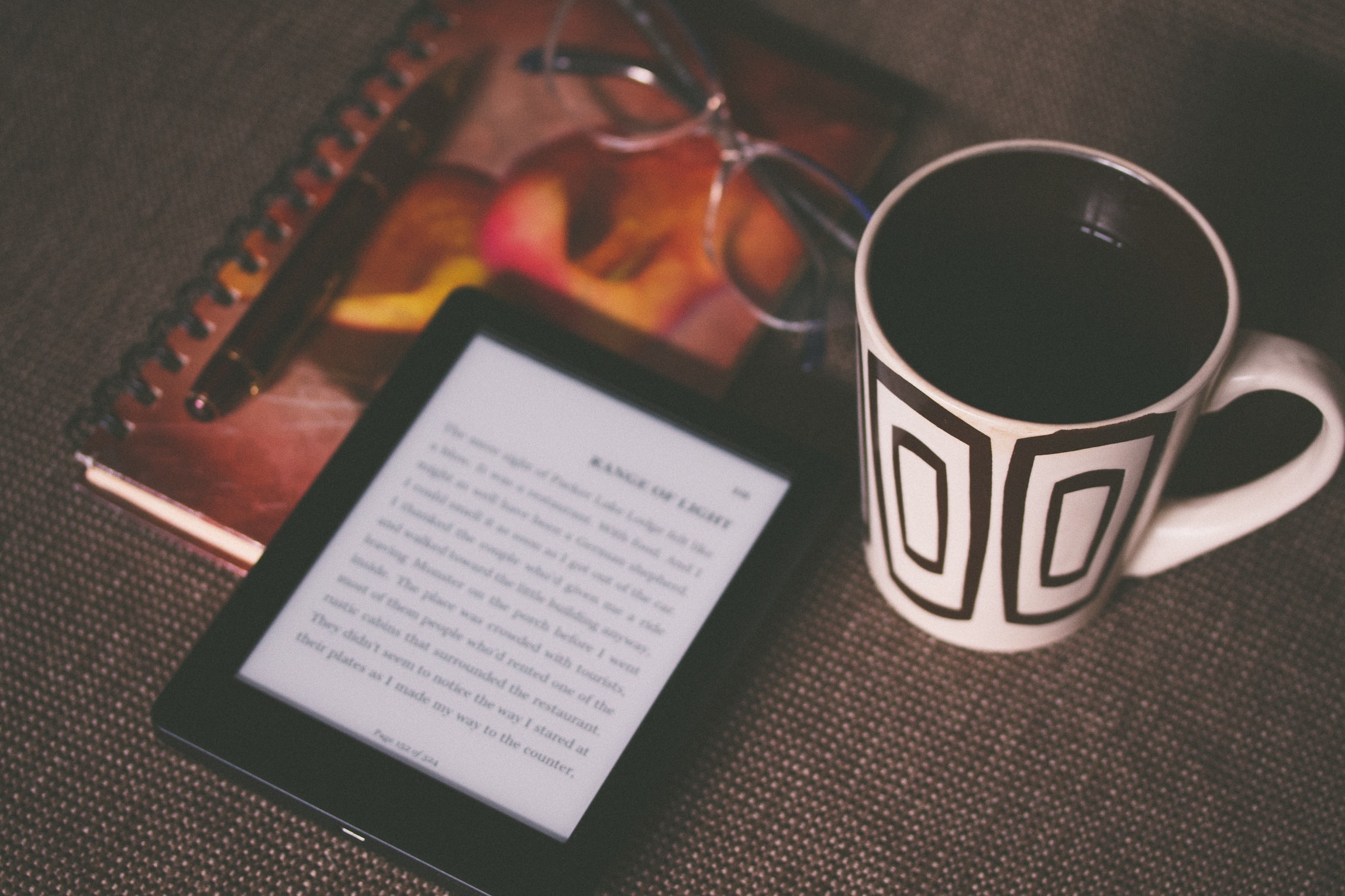 An image of an e-reader on top of a notebook and next to a cup of coffee.