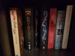 An image of a bookshelf. On the far left sits a brown and tan cookbook with "Bake Sale" written in cursive font.