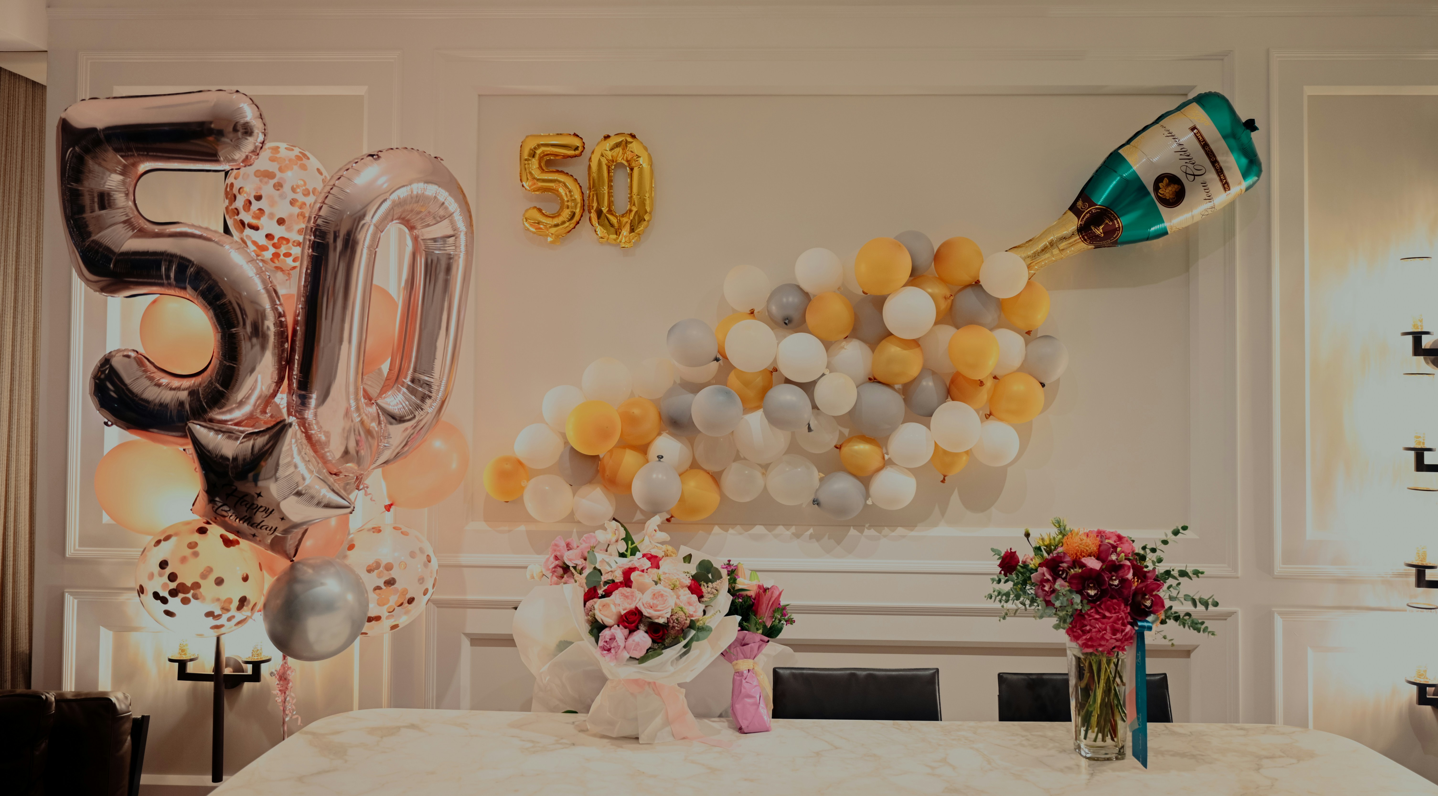 A festive room decorated for a 50th celebration with large ‘50’ balloons, a creative arrangement of smaller balloons resembling champagne bubbles, and beautiful flower bouquets on a table.