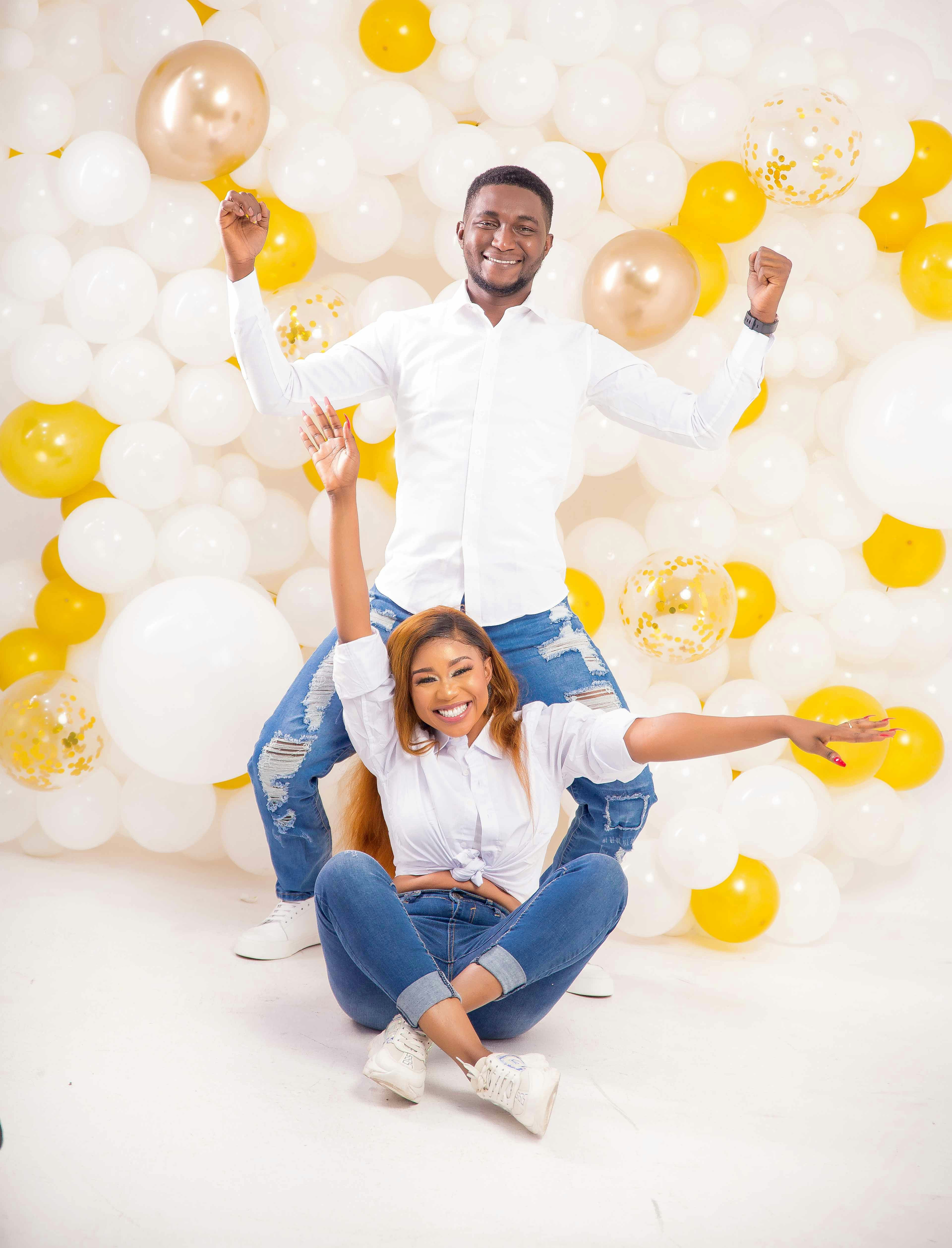 A couple posing for a photoshoot against while, yellow, and gold balloons in the background