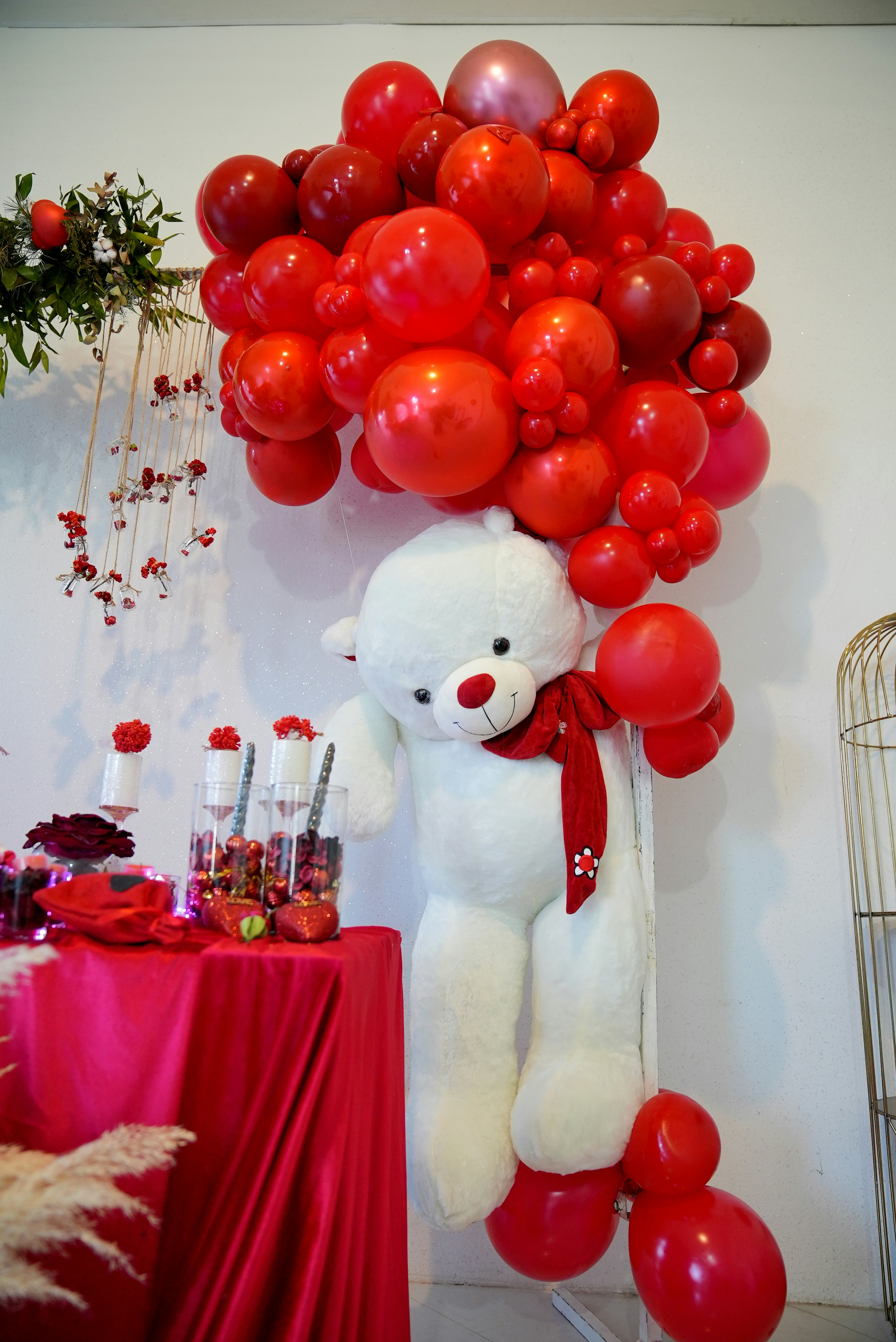 A large white teddy bear with a red bow is seated next to a table covered in a red cloth, adorned with various items including flowers and bottles. Above the bear, there is an arrangement of red balloons of different shades and sizes, creating a festive atmosphere