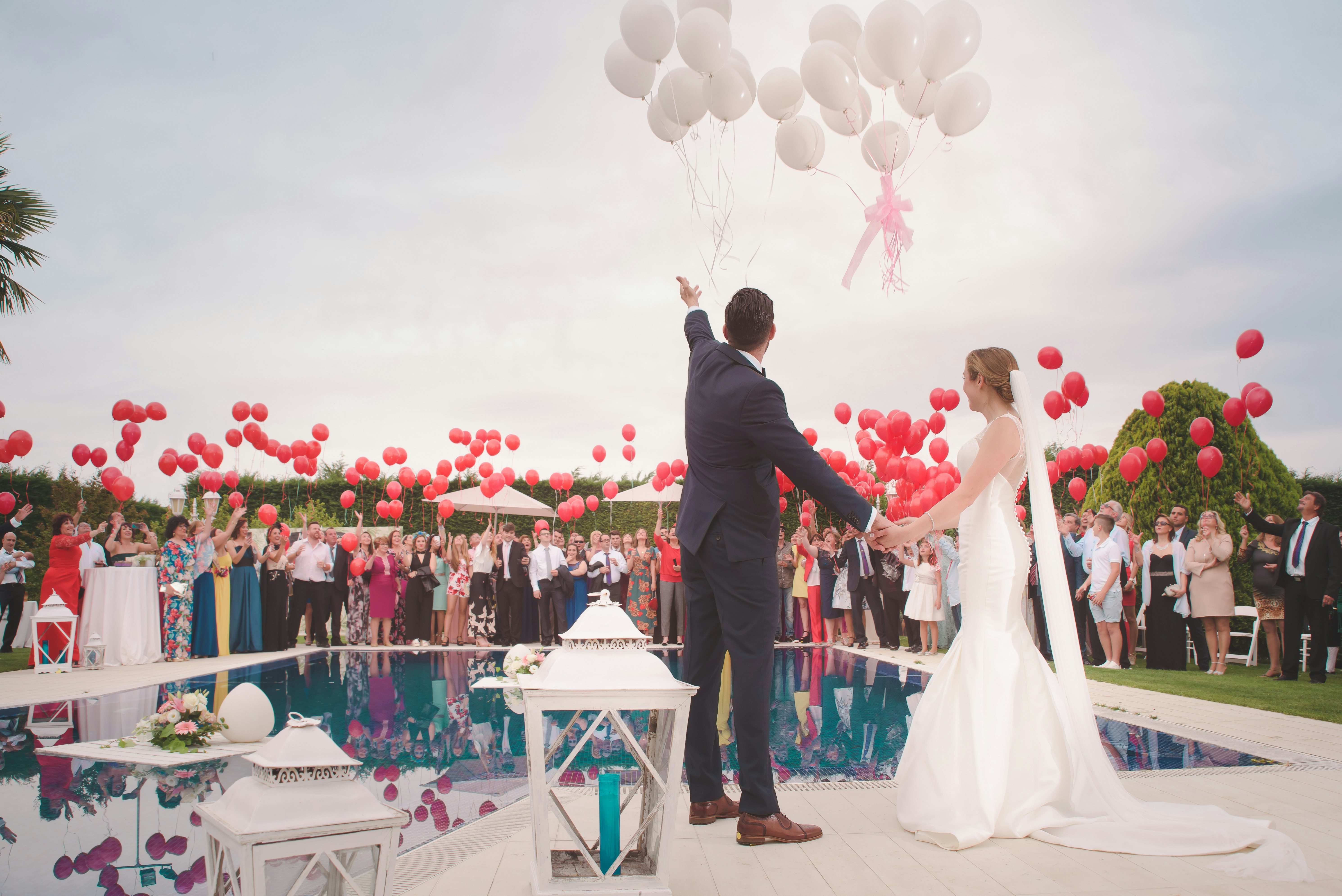 A couple releasing balloons into the sky during a wedding ceremony, surrounded by guests and standing next to a pool adorned with flower petals.