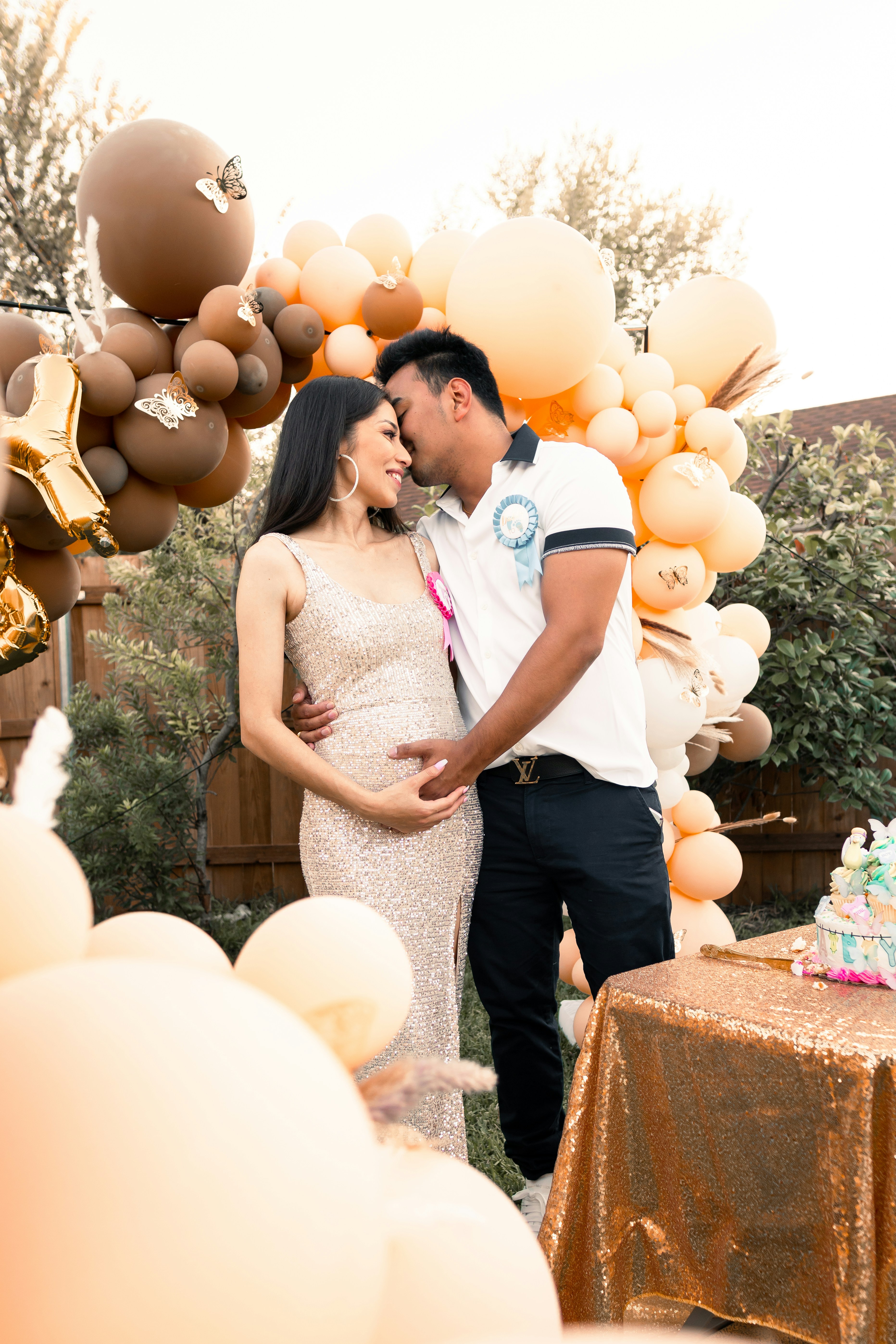 A couple embracing each other amidst a celebration, surrounded by balloons and a decorated table, in an outdoor setting.