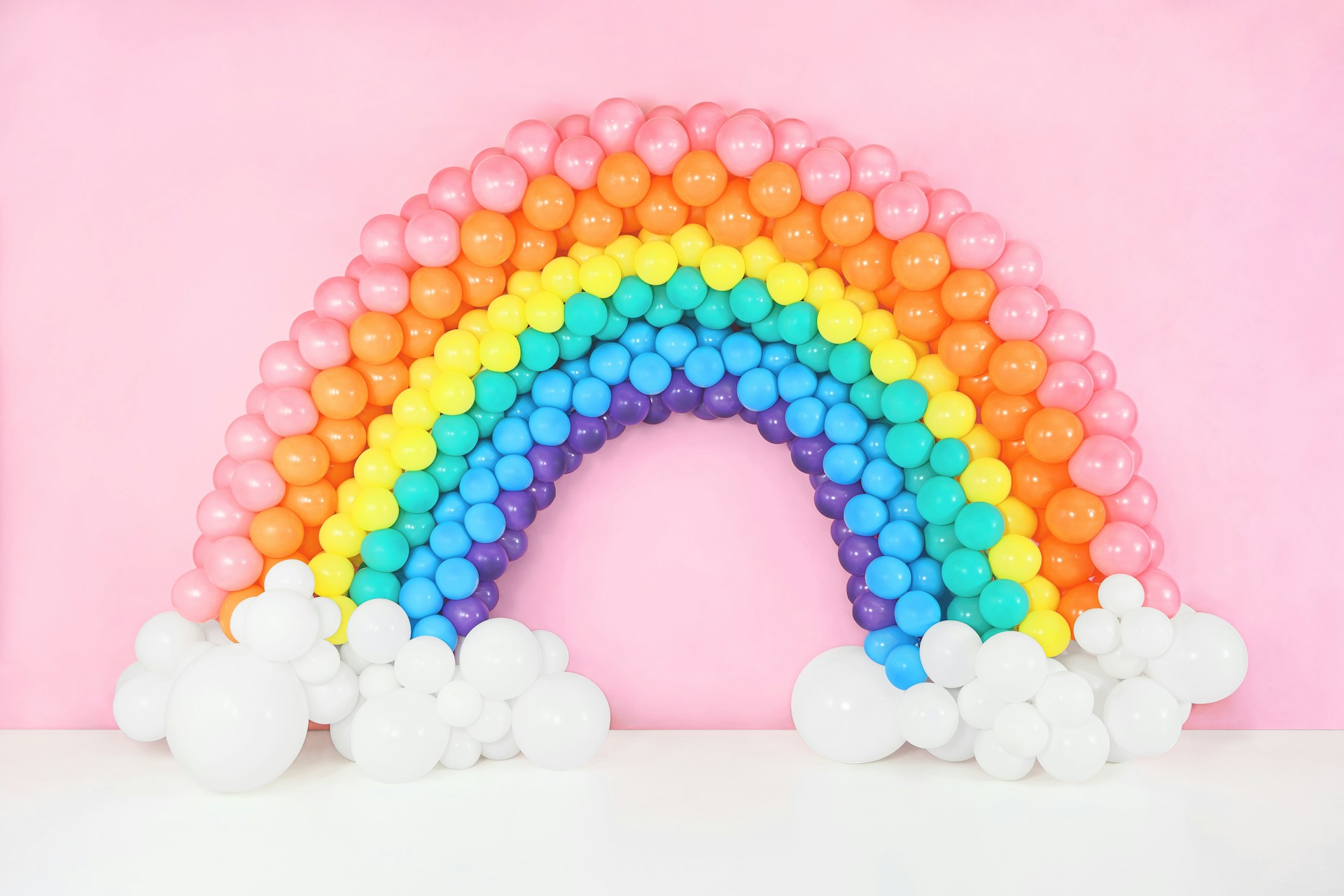 A colorful balloon rainbow with clouds, made of pink, orange, yellow, green, blue, and purple balloons against a pink background.