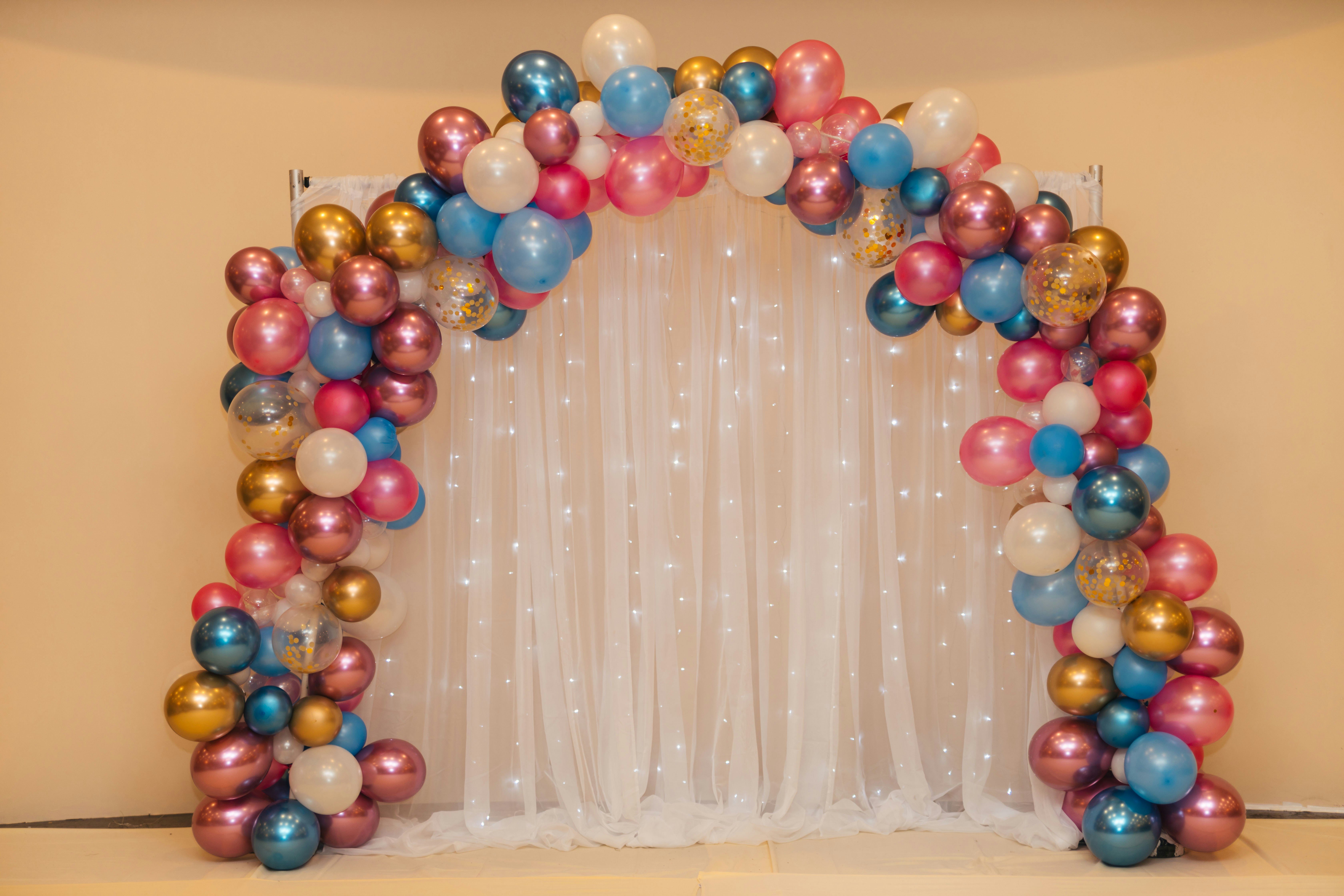 A colorful balloon arch with a mix of pink, blue, gold, and white balloons surrounding a sheer white curtain backdrop adorned with twinkling lights