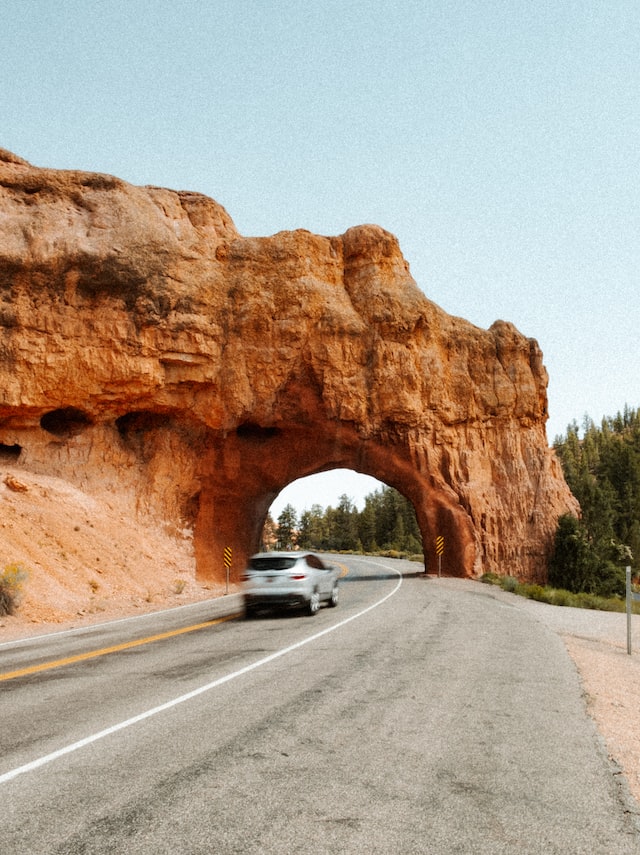 a silver car drives along under an orange rock tunnel that towers over the road.