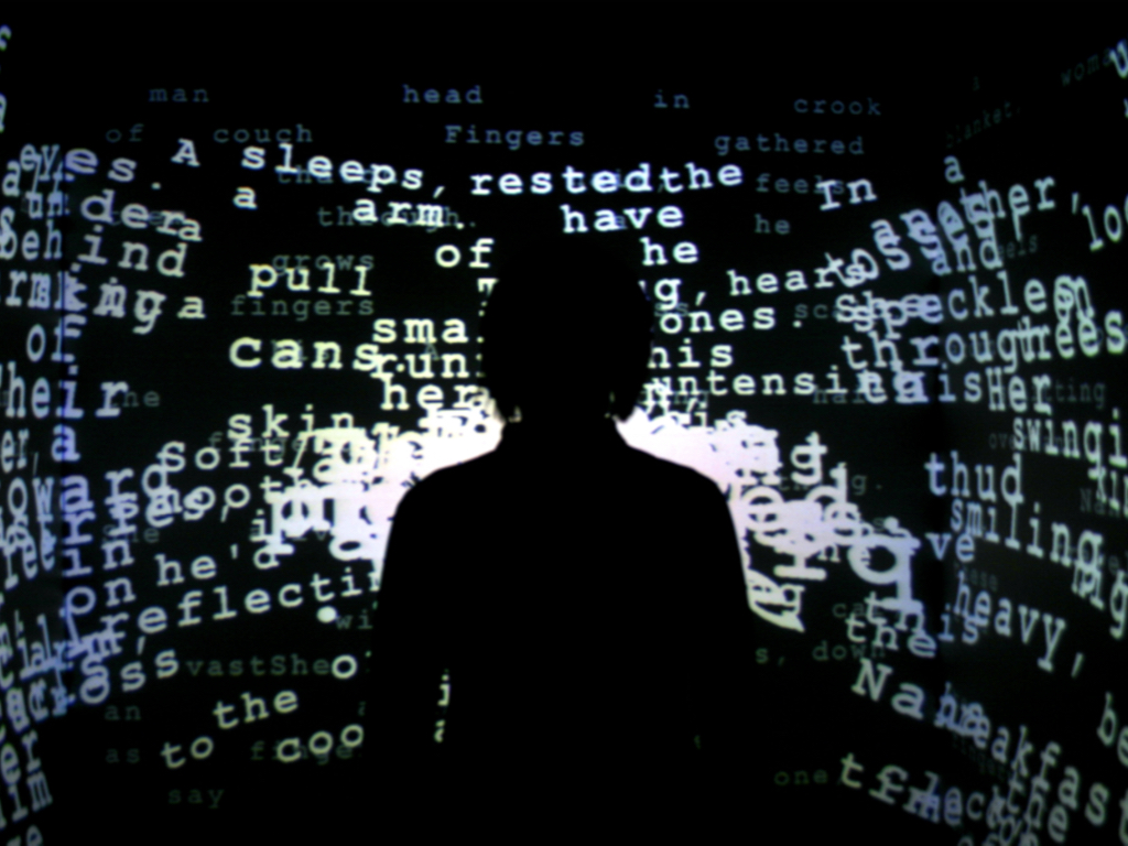 Silhouette of a person standing in front of a dark, immersive projection filled with scattered, overlapping white text fragments, creating a chaotic and poetic digital environment.