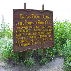Sign that reads: Laura's Dugout Home on the Banks of Plum Creek: The Charles Ingalls Family home was located here in the 1870s. This depression is all that remains since the roof caved in years ago. The prairie grasses and flowers here grow as much as they did in Laura's time. And the spring still flows nearby.