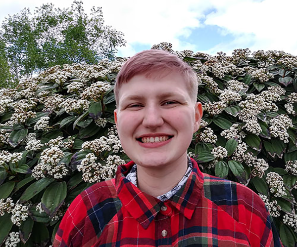 boy smiling while wearing a red flannel and standing behind a flower bush.