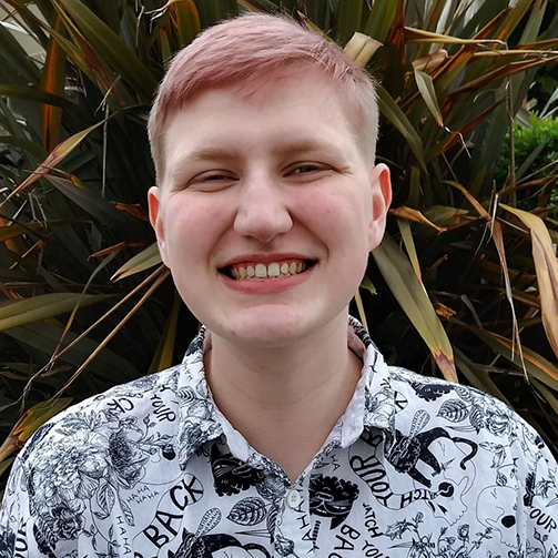 picture of man wearing a button-up shirt with a plant behind him, smiling.