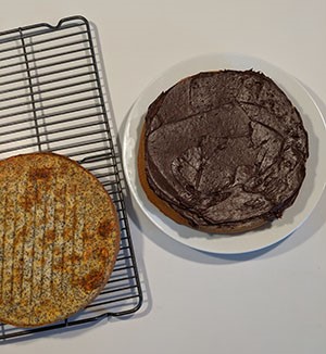 Two round cakes, one on the left is unfrosted on a cooling rack, one on the right has chocolate frosting. 