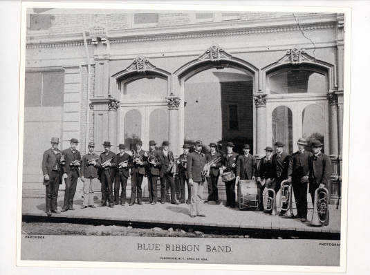 Line of men wearing uniforms, holding instruments, one man standing in front, text “Blue Ribbon Band”, grayscale photograph