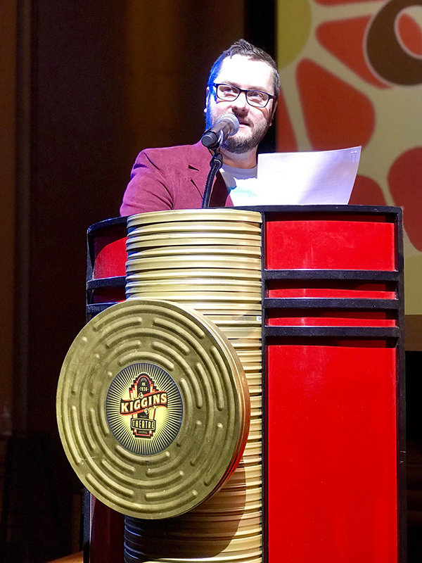 Speaker in suit standing behind red and gold podium, Kiggins Theater