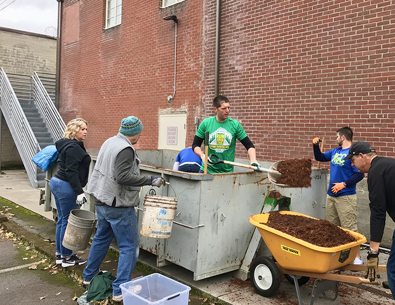 Man standing in large dumpster shoveling out dirt, others surround with buckets and wheelbarrow