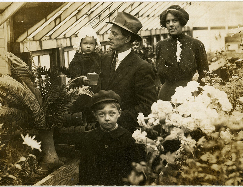 Family, child in front, man holding baby and woman standing behind surrounded by plants, vintage photograph