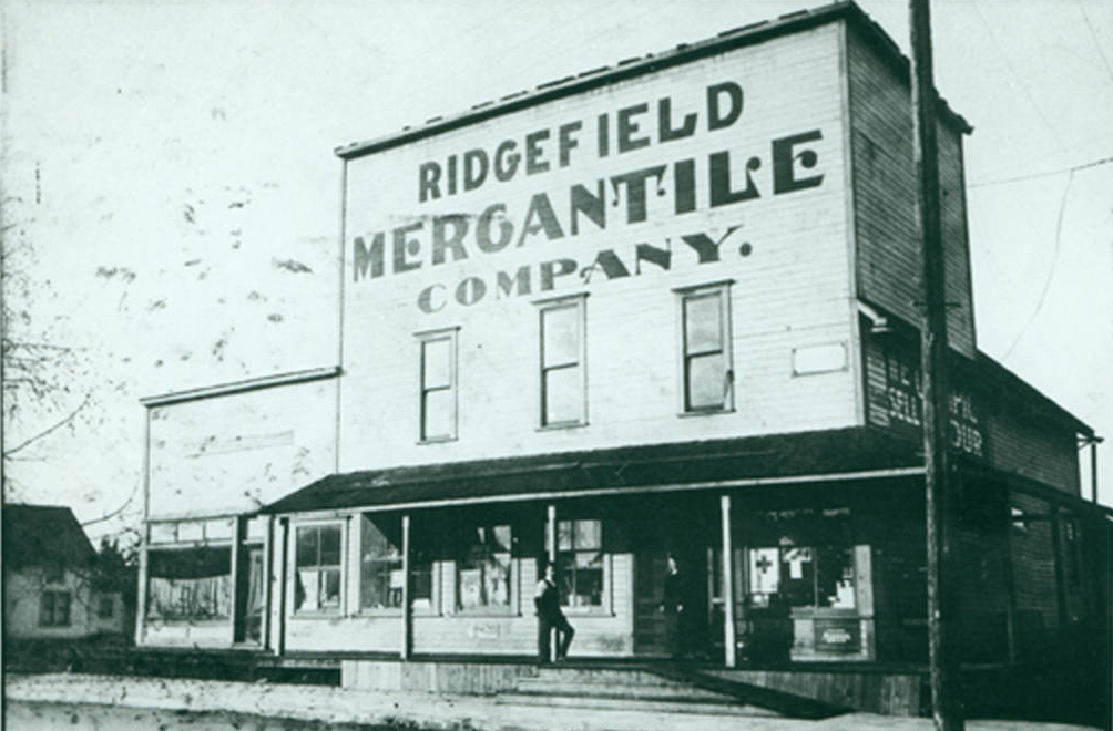 Wood building with wood awning, person standing out front, text on building reads “Ridgefield Mercantile Company”, greyscale 