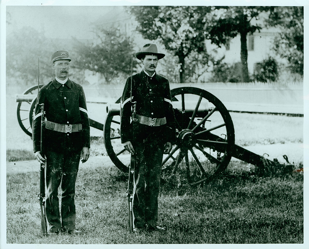 Two men in military uniform standing at attention holding muskets, cannon in background, greyscale