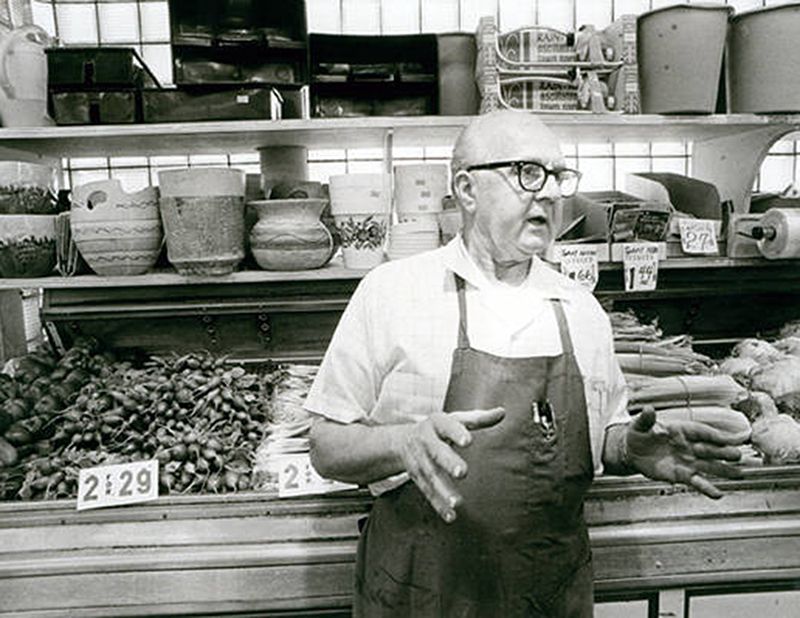 Man in apron and glasses talking produce lined behind him