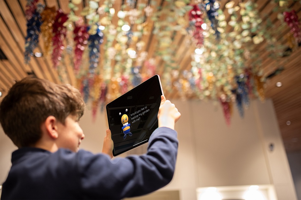 A child standing under Phonic Bloom sculpture looking at the Phonic Bloom app on a tablet with an animated bee mascot on the screen