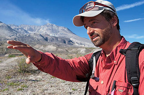 Professor of Biological Sciences John Bishop performing ecological research at Mt. St. Helens, gesturing while explaining his findings in a mountainous landscape.